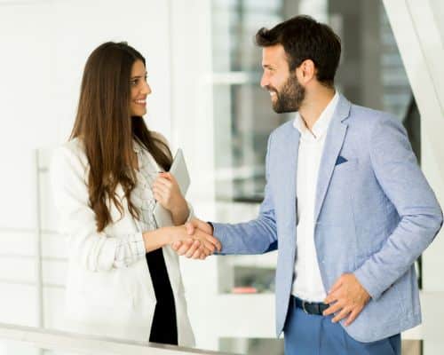 A man and a woman dressed in business attire and shaking hands while smiling. This is representative of a narcissist couple whose relationship resembles a series of business transactions. The article is about whether narcissists can be in relationship with each other.