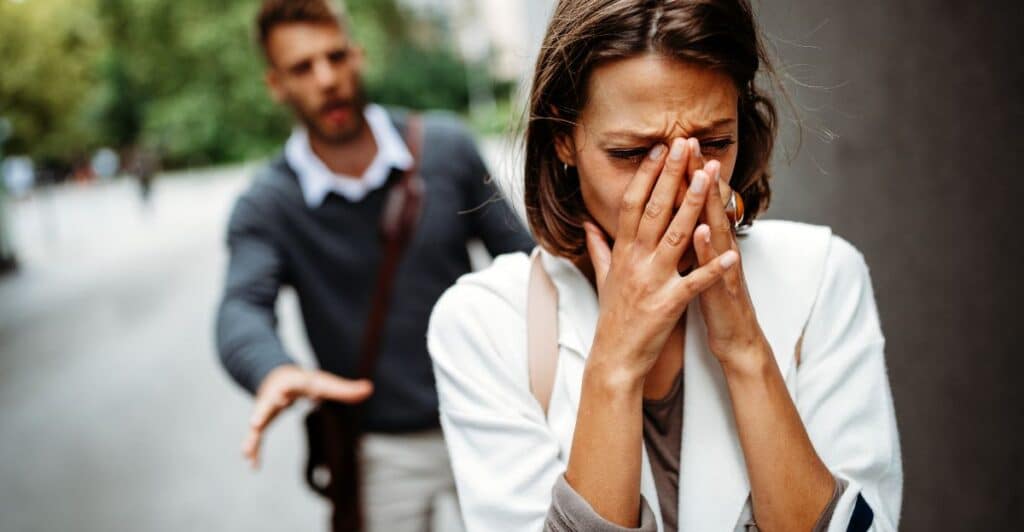 A woman covering her face with her hands to hide her distress while her husband is behind her reaching out in anger, symbolizing the title of the article, "Do Narcissists Care if You Divorce Them?"