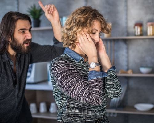 A distressed woman standing up hiding her face while her husband is yelling right behind her, signifying the name of the article, "Will a Narcissist Divorce You."