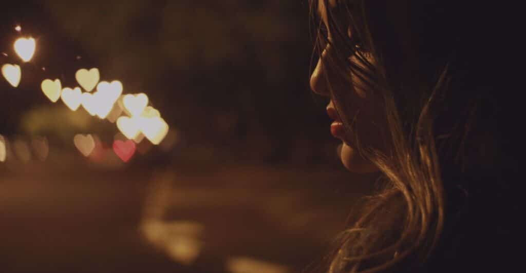 A picture of part of a woman's face on the right hand side of the picture, seemingly on a street at night. In the background on the left hand side, are street lights reflecting as hearts. The woman is sad, representing a breakup and the subject of the article, "How to Safely Leave a Narcissist for Good."