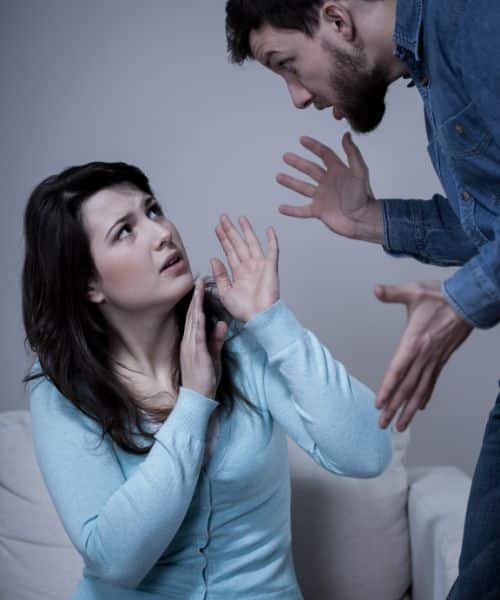 A man standing over his wife, who is cowering back with her hands blocking his, indicating the narcissist being intimidating, one of the many narcissist reactions to divorce.