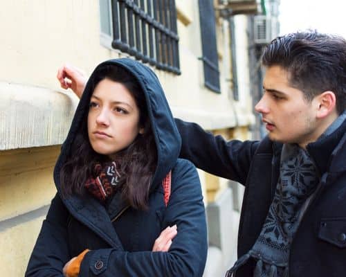 A man and woman on the street. The woman, dressed in a sophisticated dark blue jacket with hood is on the left and looking away from the man, visibly irritated. The man, on the right is wearing an elegant dark blue coat with blue and gray Nordic scarf. He is leaning his right hand on the building right behind the lady's head and trying to explain his way out of trouble. This represents the name of the article, "Covert Narcissist Checklist: Help to Find the Truth."