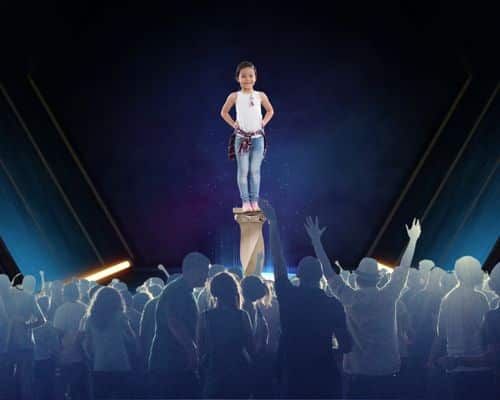 A little girl around 6 years old standing on a pedestal in front of a large crowd. This picture represents children who become narcissists because their parents and others put them on a pedestal their whole life. The name of the article is "How do Narcissists Become Narcissists."