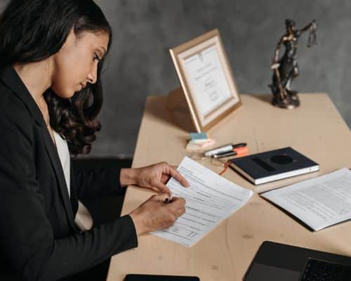 A female lawyer in a black suit at a desk doing paperwork. On her desk is her paperwork, a book/journal, a law certificate in a stand-up frame, and a small justice is blind statue. This photo represents the title of the article, "How to Expose a Narcissist in Court" and the subtopic, "To Expose a Narcissist in Court, Hire the Best Lawyer."
