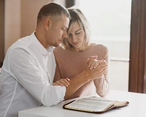 A couple sitting at a table, holding hands and praying, indicating they are praying for their marriage before it is too late. The name of the article is "When to Stop Praying for Marriage Restoration."