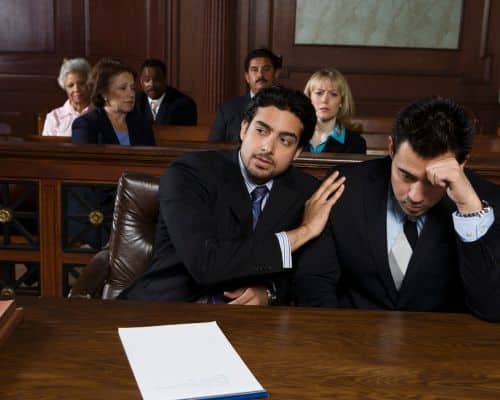 A man sitting in court at a table with his lawyer, who is patting him on the shoulder. The man has his head down, looking defeated, as though he is a victim in the courtroom. This photo represents the title of the article, "Crazy Games Narcissists Play During Divorce."