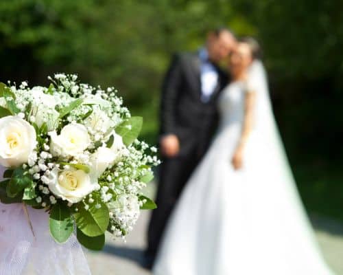 A bridal bouquet in the left foreground with a couple in wedding gown/tux in the background posing while very close to each other. This photo represents the article, "Is it a Sin to Remarry After Divorce?"