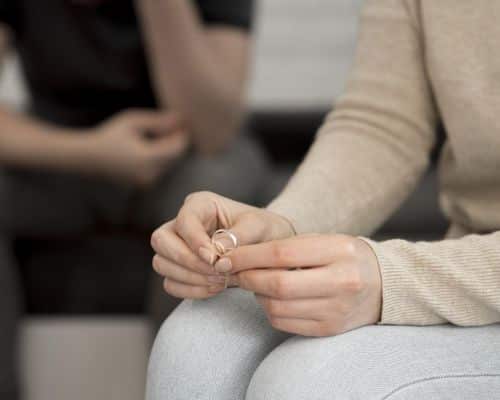A woman sitting on a sofa with just her knees and hands holding her wedding ring visible. In the background is her husband's torso, arms, and legs. both postures are sad. This photo represents the title of the article, "What it Looks Like When God Leads You to Divorce."