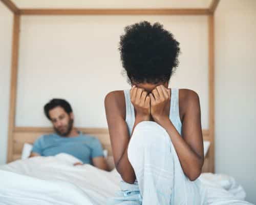 A woman sitting on a bed in the foreground, hiding her face in her fists, with her husband leaning against the headboard of the bed behind her. This photo represents the title of the article, "How Will God Protect me From a Narcissist."