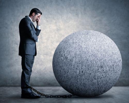 A man in a suit standing in front of a huge ball and chain with his hand covering his face. This talks about the narcissist's attachment to lifelong shame in the article, "What Happens When a Narcissist Loses in Court."