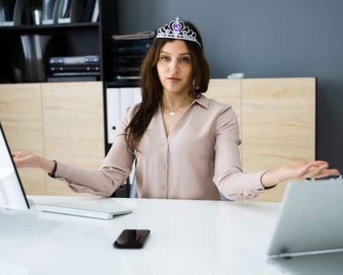 A businesswoman sitting at her desk with a crown on her head and hands out to her sides, behaving as though she is royalty. This represents an overly proud person, symbolizing the title of the article, "What Words Does the Bible Use for Narcissists/Narcissism?"