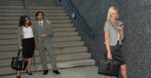 A couple in business attire at the foot of subway stairs, the woman's arm is around the man. He is looking off in the distance gazing at a woman in business attire walking past them in the foreground of the picture. He is looking at her as though he is interested in her romantically. This photo represents the title of the article, "Dealing With Your Narcissist's Wandering Eye."
