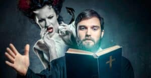 A priest in the foreground of the picture holding an open Bible up in front of him. Behind his right shoulder is a female demon, looking on in horror at what he is reading in his Bible. This photo represents the title of the article, "What are the Spiritual Roots of Narcissism."